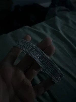 Grått broderat armband Dior - Snyggt grått armband från Christian Dior med texten 'J'ADIOR' broderad på bandet. Armbandet är gjort i tyg och har tofsar i ändarna för justerbar passform. Perfekt accessoar för att lyfta din outfit.