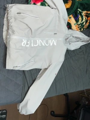 Ljusgrå vindjacka från Moncler - Snygg ljusgrå vindjacka från Moncler med huva och dragkedja framtill. Jackan har vita detaljer, stora Moncler-trycket på bröstet och två snedställda fickor med dragkedja. Materialet är glansigt och lätt, perfekt för blåsiga dagar.