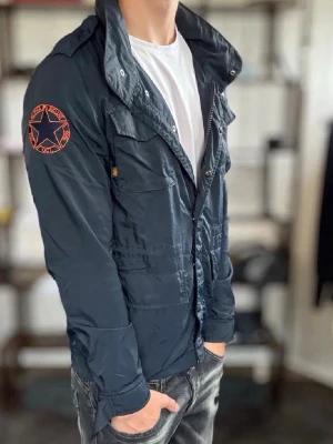 Field jacket - Snygg mörkblå fieldjacket med hög krage, dragkedja och tryckknappar. Jackan har flera fickor framtill och en orange patch med stjärna på vänster ärm. Materialet är tunt och lätt, perfekt för blåsiga dagar. Passar till en avslappnad streetstil.