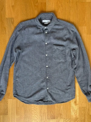 Grå Flannelskjorta från Samsøe Samsøe - Stilren grå flannel i mjuk bomull med klassisk krage och bröstficka från Samsøe Samsøe. Skjortan är gjord av 100% bomull med ett portugisiskt tyg. Skjortan är i storleken L och är i ett använt skick. Perfekt för en avslappnad men snygg look.