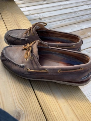 Bruna seglarskor från Sperry - Klassiska bruna seglarskor från Sperry i läder med råa mockasinsömmar och snörning runt hela skon. Skorna har vita, räfflade gummisulor med texten 'NON-MARKING' undertill. Perfekta för en avslappnad och stilren look. Storlek 44