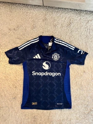Manchester United blå matchtröja - Snygg blå Manchester United matchtröja från Adidas med vita detaljer och tre ränder på axlarna. Tröjan har klubbmärke på bröstet, Snapdragon-logga och ett mönster över hela tyget. Tillverkad i lätt och ventilerande material, perfekt för träning eller match.