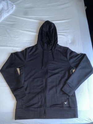 Mörkgrå tränings hoodie - Säljer en stilren mörkgrå tränings hoodie med dragkedja framtill och justerbar huva. Hoodien har långa ärmar, ribbade muddar och två fickor framtill. Liten vit logga nertill på ena sidan. Perfekt för en avslappnad look.