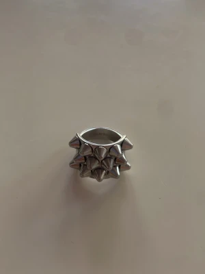 Silvrig ring med nitar - Cool ring i silverfärgad metall med flera spetsiga nitar runt hela ringen. Ringen har en chunky och edgy design som verkligen sticker ut och passar perfekt för dig som gillar statement-smycken.