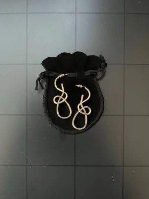 Guldiga örhängen i knutdesign - Säljer ett par unika örhängen i guldtonad metall med en cool knutformad design. Örhängena har en droppformad siluett och stift för enkel användning. Perfekta för dig som gillar statement-accessoarer och vill sticka ut med något annorlunda.