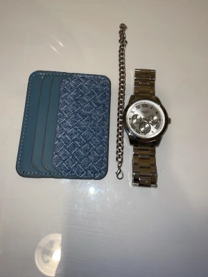 Blå kortfodral i konstläder,silver armband och silver klocka. - Snyggt blått kortfodral med fyra fack, tillverkat i konstläder och tyg med ett rutigt mönster framtill. Perfekt för att hålla koll på dina kort i fickan eller väskan. Stilren design med vita sömmar och modern känsla. Men en liten touch klocka och armband😃