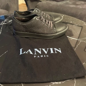 Svarta sneakers från Lanvin - Snygga svarta sneakers från Lanvin i mycket bra skick. De har en stilren design med snörning och en kombination av skinn och textilmaterial. Perfekta för både vardag och festliga tillfällen. Sulan är i bra skick och ger bra grepp.