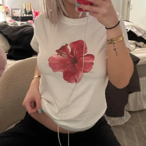 Vit t-shirt med röd blomma - Säljer en superfin vit t-shirt med en stor röd blomma på framsidan. Perfekt för sommardagar eller när du vill ha något enkelt men ändå snyggt. T-shirten har en normal passform och är kortärmad. Den är i bra skick och gjord av mjuk bomull. Passar perfekt till jeans eller shorts!