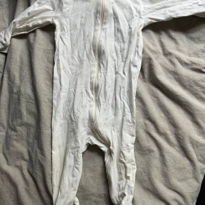 Vit onesie från Kappahl - Säljer en söt vit onesie från Kappahl i storlek 80, passar 9-12 månader. Den har en dragkedja framtill för enkel påklädning. Perfekt för mysiga dagar hemma eller som pyjamas. 🍼