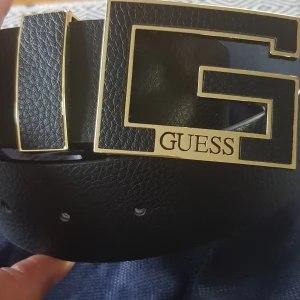 Svart skärp från Guess med gulddetaljer - Säljer ett snyggt svart skärp från Guess i skinn med en stor, fyrkantig guldfärgad spänne. Perfekt för att ge en lyxig touch till din outfit. Skärpet har flera hål för justerbar passform. Använd det till fest eller vardag för en stilren look.