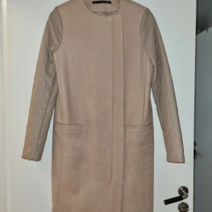 Beige kappa från Annarita N - Säljer en elegant beige kappa från Annarita N i storlek 42 men motsvarar M. Den har quiltade ärmar och en stilren dragkedja framtill. Perfekt för höst och vår, med en klassisk och tidlös design. Passar både till vardags och festliga tillfällen.