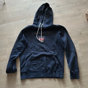 Mörkblå hoodie från GANT - Säljer en snygg mörkblå hoodie från GANT med deras klassiska logga och ett coolt emblem på bröstet. Strlk M