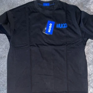 Svart t-shirt från Hugo - Säljer en svart t-shirt från Hugo i nytt skick. Den har en cool blå logga på bröstet som verkligen poppar. Perfekt för både vardag och fest! T-shirten är kortärmad och gjord i mjuk bomull för bästa komfort. Passar perfekt till jeans eller shorts.