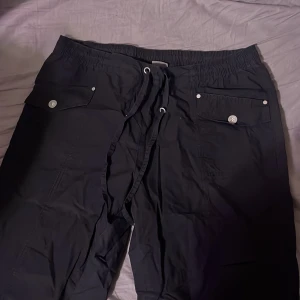Svarta byxor med snörning - Säljer ett par svarta byxor i bra skick. De har en bekväm elastisk midja med snörning och praktiska fickor med knappar. Perfekta för en avslappnad stil eller vardagsbruk. Passar till det mesta och är superbekväma!