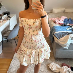 Blommig offshoulder klänning - Säljer en superfin blommig offshoulder klänning i vitt med rosa och gröna blommor. Den har en söt volangdetalj vid kjolen och är perfekt för sommarens fester. Klänningen är i mycket bra skick. Perfekt för en solig dag eller en sommarkväll! 🌸