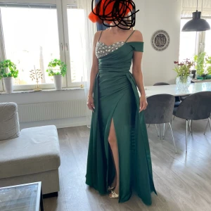 Grön långklänning med paljetter balklänning bröllop - Säljer en elegant grön långklänning med paljettprydd överdel och offshoulder-design. Klänningen har en smickrande slits framtill och är perfekt för festliga tillfällen. Den är i mycket bra skick och ger en glamorös look.