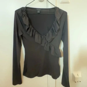 Svart långärmad topp med volang - Snygg svart långärmad topp  med volangdetaljer längs halsringningen. Perfekt för både vardag och fest. Toppen har en figurnära passform och är gjord i ett mjukt material som känns skönt mot huden.