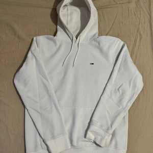 Vit hoodie från Tommy Hilfiger (Tommy Jeans) - Säljer en stilren vit hoodie från Tommy Jeans i storlek M. Den har en klassisk design med en liten logga på bröstet och justerbar huva med snören. Perfekt för en avslappnad look eller kyliga dagar. Skön och mjuk kvalitet!