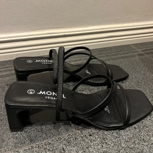 Svarta sandaler med klack från Monki - Snygga svarta sandaler från Monki i veganskt material. De har en stilren design med smala remmar och en fyrkantig tå. Perfekta för både vardag och fest! Klacken ger en elegant touch utan att vara för hög.