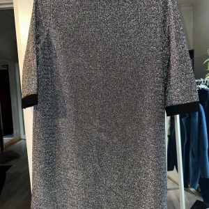 Glittrig silverklänning - Snygg glittrig silverklänning med trekvartsärm. Perfekt för festliga tillfällen! Klänningen har en rak passform och är i ett glittrande material som verkligen fångar ljuset. Svarta detaljer vid ärmsluten ger en fin kontrast.