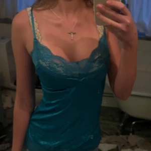 Turkos linne med spetsdetaljer - Säljer ett snyggt turkost linne med spetsdetaljer vid urringningen. Linnet har smala axelband och en figurnära passform, perfekt för sommardagar eller en utekväll. Det är i bra skick och ger en fräsch och stilren look.