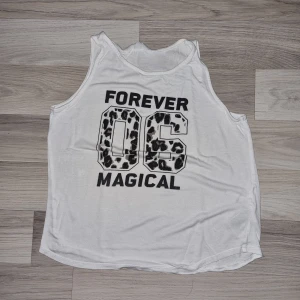 Vit linne från H&M - Säljer en vit linne från H&M med texten 'FOREVER MAGICAL 06' i leopardmönster. Perfekt för sommardagar eller som ett lager under en jacka. Toppen är i bra skick och passar både till vardags och avslappnade tillfällen.