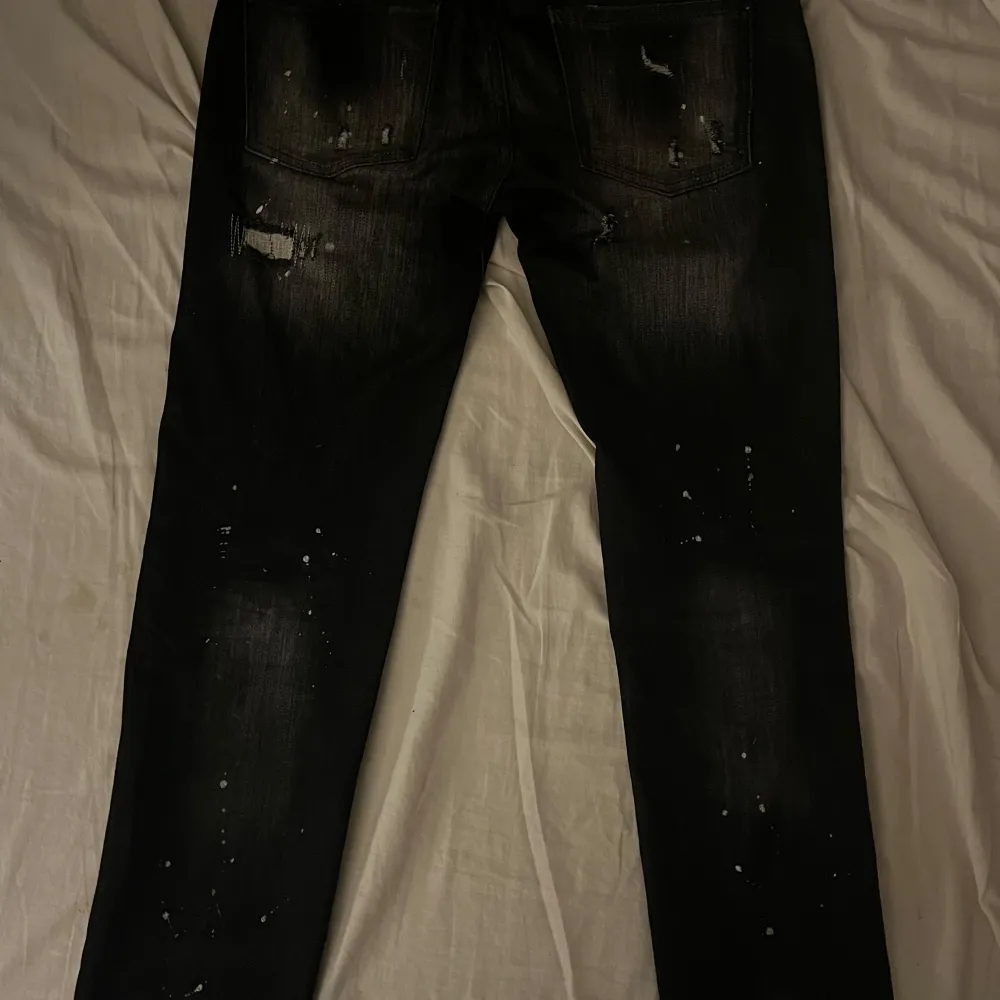 Säljer ett par svarta jeans från Dsquared2 i storlek 48. De har en cool distressed look med slitningar och färgstänk som ger en edgy stil. Perfekta för en avslappnad men trendig outfit.. Jeans & Byxor.