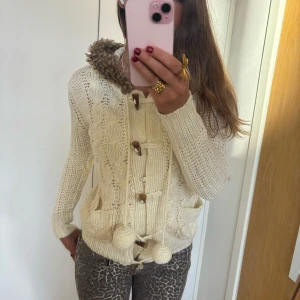 Beige stickad kofta med pälskantad huva - Mysig beige stickad kofta med pälskantad huva och träknappar. Inte stickig. Perfekt för kyliga dagar med sin varma och bekväma design. Den har långa ärmar och dekorativa tofsar framtill. Passar perfekt till höst- och vintergarderoben! jag är 168 och det är mina bilder, köp via köp nu direkt eller Swish, kvar tills markerad som såld ❤️