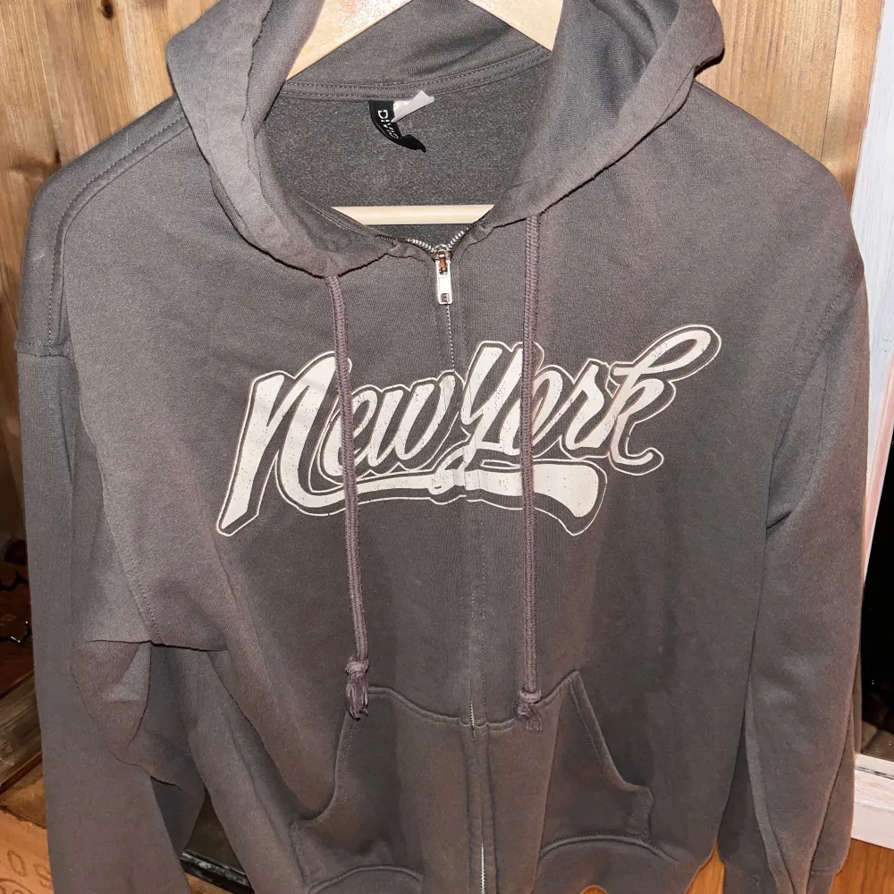 Säljer en grå hoodie från Divided med ett coolt New York-tryck på framsidan. Den har dragkedja och en skön huva med snören. Perfekt för en avslappnad stil eller mysiga dagar hemma. Passar bra till jeans eller leggings!. Hoodies.