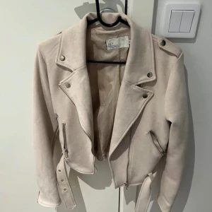 Beige mockajacka från New Trendy - Säljer en snygg beige mockajacka från New Trendy. Jackan har en cool bikerstil med dragkedjor och ett bälte i midjan. Perfekt för höst och vår! Den är i mycket bra skick och har långa ärmar. Passar perfekt för en trendig look. Använd 2-3 gånger.