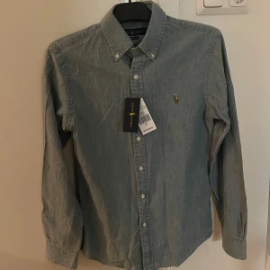 Helt ny Ralph Lauren skjorta - Säljer en snygg blå skjorta från Ralph Lauren i nytt skick. Skjortan har en klassisk krage och knappar framtill. Den är långärmad och har det ikoniska broderade logotypen på bröstet. Skriv om ni har frågor!💕