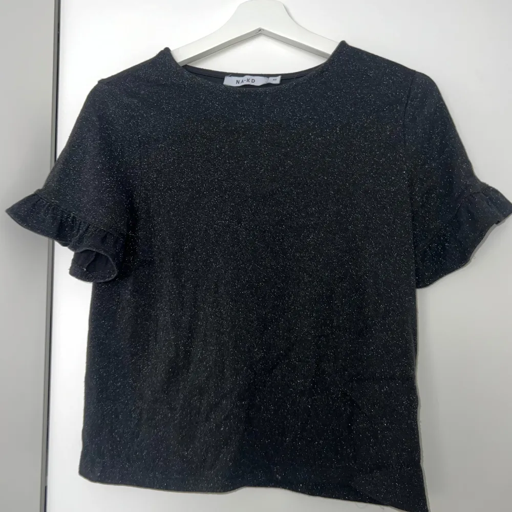 Svart glittrig topp från NA-KD i storlek XS. Korta ärmar med volangdetaljer. Älskar denna men tyvärr för liten för mig 🥹🪩. Yläosat.
