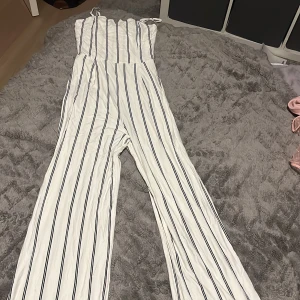 Vit och svart randig byxdress från Bershka - Säljer en superfin vit byxdress från Bershka med svarta ränder. Den har en smickrande passform med vida ben och smala axelband. Perfekt för sommardagar eller en avslappnad festkväll. Använd ett fåtal gånger, så den är i mycket bra skick. Passa på att fynda! Den har en super super liten fläck på insidan som jag visade på bild 4, absolut ingenting man märker av eller ser dock!🥰