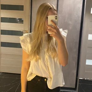 Vit blus med volangdetaljer - Säljer en söt vit blus med volangdetaljer på ärmarna. Blusen har en rund halsringning och fina spetsdetaljer framtill. Perfekt för vår och sommar, både till vardags och fest. Den är i bra skick och passar till många olika outfits!