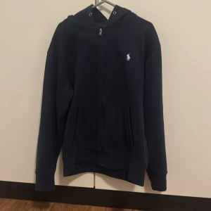 Mörkblå hoodie från Ralph Lauren - Säljer en snygg mörkblå hoodie från Ralph Lauren i storlek M. Den har en dragkedja framtill och en klassisk logga på bröstet. Perfekt för både höst och vår, och den är superbekväm att ha på sig. Passar till alla tillfällen! 💙