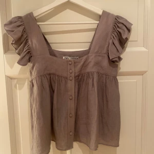 Topp från Zara - Säljer en söt grå topp från Zara med volangdetaljer på axlarna och knappar framtill. Passar både till vardags och festliga tillfällen. Storlek S. Aldrig använd! 