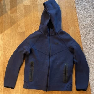Mörkblå hoodie från Nike Tech - Säljer en mörkblå hoodie från Nike med dragkedja och huva. Jackan har långa ärmar och två fickor med dragkedjor framtill. Perfekt för en sportig look eller en avslappnad dag.str: 147/158