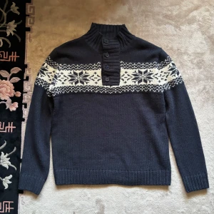 Mörkblå stickad tröja med mönster - Säljer en mörkblå stickad tröja med ett klassiskt vitt mönster över bröstet. Tröjan har en hög krage med knappar och långa ärmar. Perfekt för kyliga dagar!