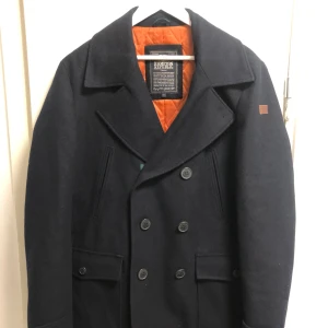 Mörkblå skepparkavaj från Hampton Republic - Säljer en stilren mörkblå skepparkavaj från Hampton Republic. Jackan har en dubbelknäppt design med stora knappar och en quiltad orange insida. Perfekt för kyligare dagar med sin klassiska look och varma foder. Nyskick då den knappt varit använd då den var för stor. 