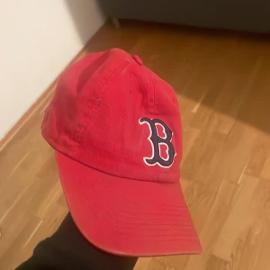 Röd keps med broderad bokstav - Snygg röd keps med en broderad bokstav på framsidan. Perfekt för en sportig look eller som en stilren accessoar. Märket är synligt på sidan av kepsen.