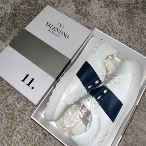 Valentino Garavani sneakers - Snygga vita Valentino Garavani sneakers med en bred mörkblå rem över sidan. Skorna har en stilren design med snörning och en bekväm sula. Perfekta för en trendig look. Helt ny aldrig använt satt bara lite tajt