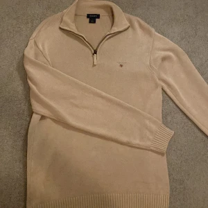Beige stickad tröja från GANT - Säljer en stilren beige stickad tröja från GANT med dragkedja vid halsen. Tröjan har ett broderat GANT-emblem på bröstet och ribbade muddar vid ärmslut och nederkant. Perfekt för en avslappnad och snygg look.