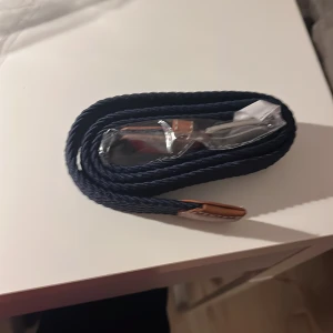 Blått flätat bälte med brunt läder -  Helt nytt Snyggt blått flätat bälte med detaljer i brunt läder och ett silverfärgat spänne. Perfekt för att ge en stilren touch till din outfit. Bältet är fortfarande i sin förpackning.