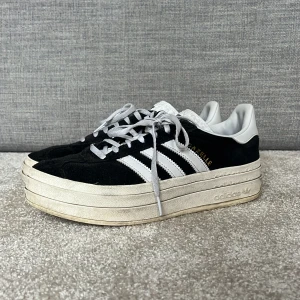 Svart Adidas Gazelle med platåsula - Snygga svarta Adidas Gazelle med vit platåsula och klassiska tre ränder i vitt på sidan. Skorna har snörning och är tillverkade i mocka med vita detaljer. Perfekta för dig som vill ha en stilren look med lite extra höjd.