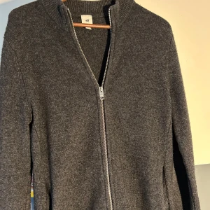 Grå kofta från H&M - Säljer en stilren grå kofta från H&Ms premium kollektion med dragkedja framtill. Koftan har en ribbad krage och långa ärma. Passar bra till både vardag och mer avslappnade tillfällen.