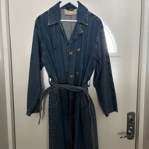 Jeanskappa från JXX - Snygg jeanskappa från JXX med knappar och bälte i midjan. Den har en klassisk krage och långa ärmar. Perfekt för en avslappnad stil med en touch av denim.