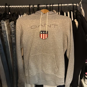 Grå hoodie från GANT - Säljer en stilren grå hoodie från GANT med logotyp och emblem på bröstet. Tröjan har en justerbar huva med vita snören och en praktisk magficka. Perfekt för både tjej och kille. Storlek S och mycket bra skick