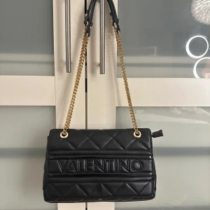 Svart väska från Valentino Mario - Snygg svart väska från Valentino Mario med quiltad design och guldfärgade kedjedetaljer. Väskan har en praktisk dragkedja och rymligt inre för dina nödvändigheter. Perfekt för att ge din outfit en lyxig touch.