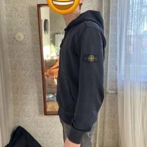 Svart hoodie med dragkedja - Säljer en marinblå hoodie från Stone Island med dragkedja och huva. Tröjan har en klassisk design med ribbade muddar och en ficka framtill. Perfekt för en avslappnad stil.