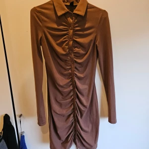 Klänning - Snygg brun klänning med långa ärmar och knappar framtill. Den har en figursydd passform med rynkdetaljer längs knappraden som ger en elegant look. Klänningen slutar strax ovanför knäna.
